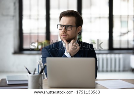 Pensive young Caucasian male boss or CEO in glasses sit at desk in office look in distance thinking or pondering, thoughtful businessman in spectacles plan or consider future business idea or success Royalty-Free Stock Photo #1751133563