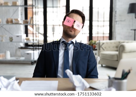 Exhausted male employee sit at desk with stickers on eyes fall asleep nap at workplace, tired businessman or boss sleeping in office, overwhelmed with work, feel fatigue exhaustion, overwork concept Royalty-Free Stock Photo #1751133602