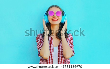 Portrait of cheerful woman in wireless headphones listening to music wearing a pink sunglasses on colorful blue background