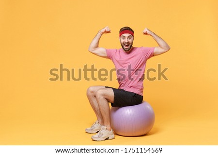 Excited young bearded fitness sporty guy sportsman in headband t-shirt in home gym isolated on yellow background. Workout sport motivation lifestyle concept. Sit on fitball, showing biceps, muscles