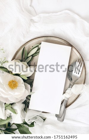 Festive summer wedding scene. Marble table setting with cutlery, olive branches, white peony flowers, stoneware plate and silk ribbon. Blank restaurant menu card mockup. Flat lay, top view, vertical.