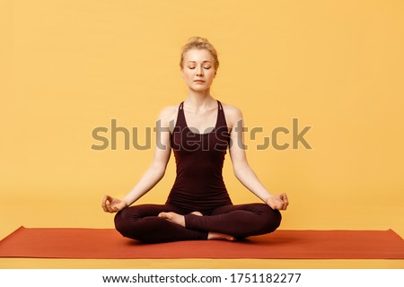 Young blond woman meditating, doing yoga lotus padmasana pose and asana. Fitness girl enjoying yoga indoors in sport clothes on yellow background, isolated. Stress-free yogic meditation practice