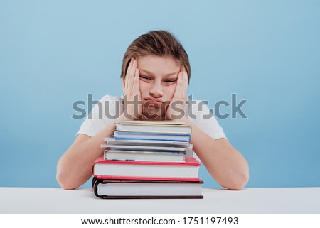 Weary diligent pupil in casual clothes looking at camera with sadness and fatigue while sitting at desk with stack of textbooks and, copybooks and leaning on hands against light blue background