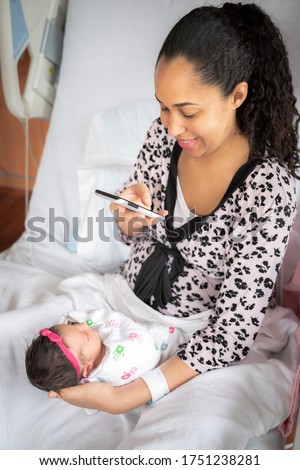 A mother in a pink animal print bathrobe sits on a hospital bed holding her newborn baby girl swaddled in a blanket while taking a photo with her mobile cell phone to share with family and friends.
