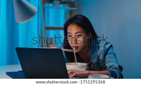 Asia freelance smart business women eating instant noodles while working on laptop in living room at home at night. Happy young Asian girl sitting on desk work overtime, enjoy relax time. Royalty-Free Stock Photo #1751295368