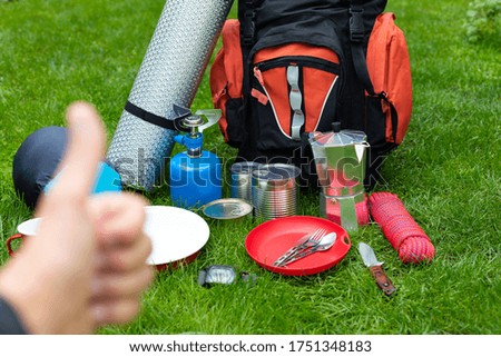 Picture of camping tools on the grass - backpack, tent, gas tank, cans, compass, etc., tourist showing thumbs up