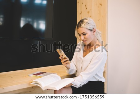 Happy young businesswoman taking photo of book pages on smartphone while sitting during work at wooden counter in modern conference room