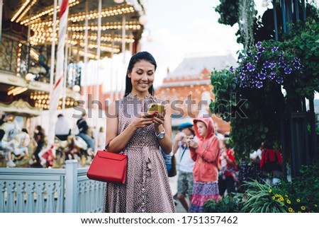 Happy ethnic female in casual outfit traveler smiling and looking at smartphone during walk near illuminated carousel in city