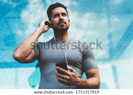 Caucasian sportive blogger with smartphone gadget in hand thoughtful looking away preparing to morning workout, muscular athletic man in electronic earphones listening mobile motivation podcast
