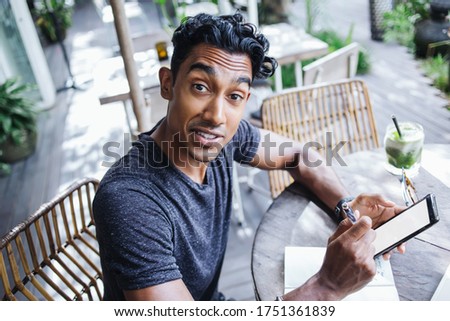 From above astonished adult Hispanic male with raised eyebrows in casual clothes holding pen and interacting with smartphone while sitting at table with notepad and green drink and looking at camera