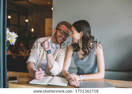 Man with pen and open notebook friendly nudging young female newbie employee while sitting together at table with laptop and laughing Royalty-Free Stock Photo #1751362718