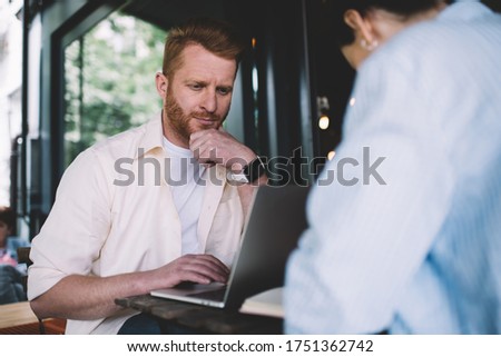 Young thoughtful redhead guy rubbing chin while using laptop for work sitting at table with crop colleague and rubbing chin in thoughts