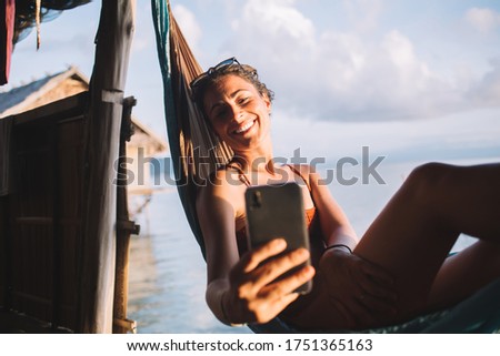 Adult joyful female in orange swimwear taking selfie on smartphone while relaxing in hammock by blue sea during beautiful sundown