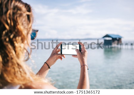 Crop back view of woman using mobile photo with white screen while picturing beautiful marine landscape during summer vacation at sea