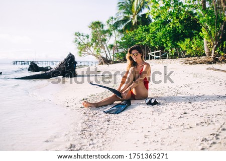 Young smiling brunette in sunglasses and red swimsuit putting on flippers on beach while sitting on sand with goggles against background of palm trees and pier