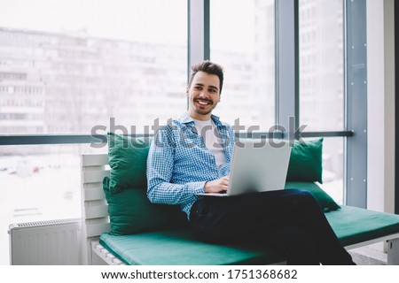 Cheerful male freelancer with modern laptop device sitting at comfortable couch and smiling, happy Caucasian IT professional in smart casual wear waiting for netbook update for working in online