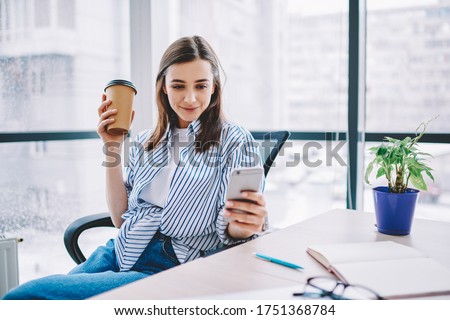 Youthful Caucasian employee in smart casual clothing using cellular application for social networking during coffee break in office interior, millennial woman sending sms during mobile messaging
