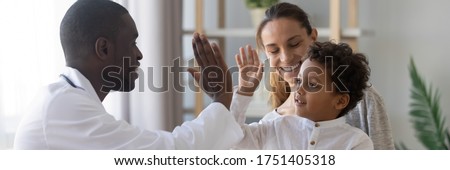 Little boy sit on mom laps give high five to African doc in white coat during visit in clinic. Horizontal photo banner for website header design, general medical check up, children healthcare concept