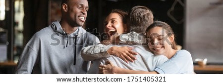Gathered in cafe diverse friends meet guy mate cuddle him feel happy young people glad to see each other. Warm friendly relations, friendship concept. Horizontal photo banner for website header design Royalty-Free Stock Photo #1751405321