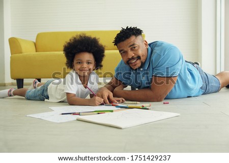 Black African family dad son together at home. Young father and little boy smile at camera and lying on warm floor and drawing coloring on book with color pencil crayon at living room