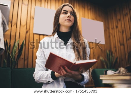 Focused young female in medical coat learning professional literature while sitting at creative room with wooden walls and plants and looking away