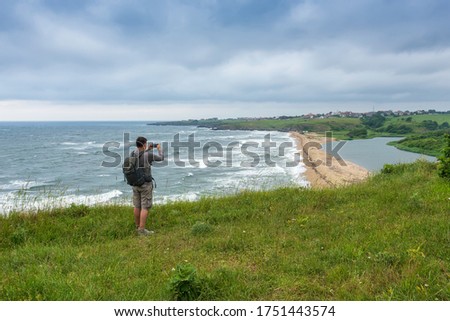 Rear view of a man taking pictures on his smart phone while standing at the edge of a rock looking toward beautiful and wild beach at the Black Sea coast and the estuary of Veleka river, Bulgaria.