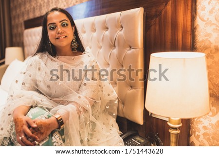 A beautiful Indian bride in traditional wear waiting in Hotel room next to lamp. A smiling girl wearing jewellery and designer Indian wear sitting close to a warm light.