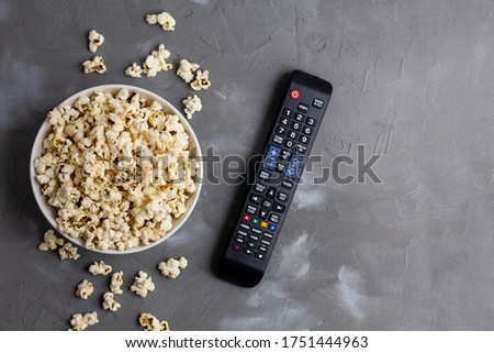 A white bowl of popcorn and TV remote on a grey background. The concept of watching TV, film, TV series, sports, shows.