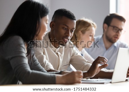 African and asian different age colleagues seated at desk in co-working room look at laptop screen discuss new corporate app, more experienced mate explain program software to intern mentoring concept Royalty-Free Stock Photo #1751484332