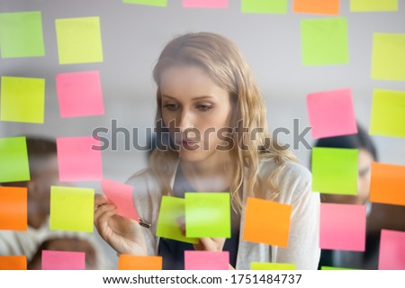 View through glass young businesswoman attaching to wall colorful post-it lot of stickers with tasks to-do list. Female member of creative department working on fresh ideas creating startup concept
