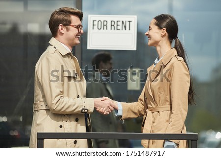 Side view waist up portrait of smiling real estate agent shaking hands with client outdoors while standing next to FOR RENT sign