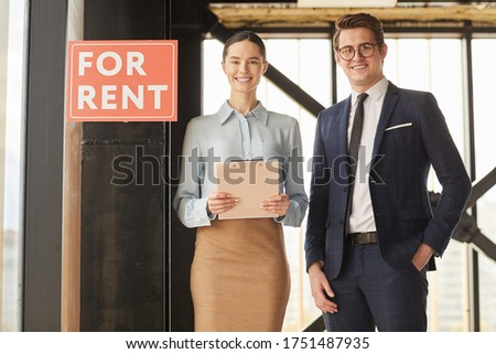 Waist up portrait of two real estate agents smiling at camera while standing next to red FOR RENT sign, copy space