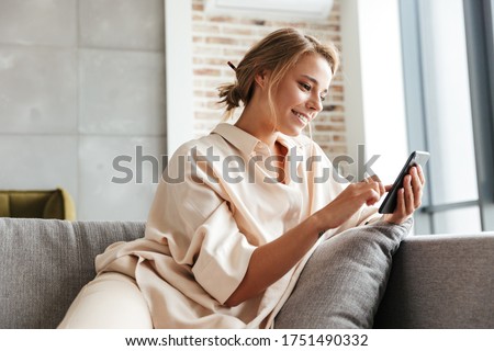 Image of happy nice woman in pajamas smiling and using cellphone while sitting on sofa at living room Royalty-Free Stock Photo #1751490332