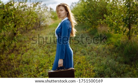 A girl in a blue dress with an old suitcase looks into the frame. The dress is strict and long. The woman's hair flutters in the wind. Around young trees and blooming Apple trees. High quality photo