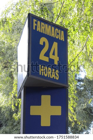 Poster with the sign, translation: "24 hour pharmacy" on the sidewalk. Street sign in blue with letters and yellow cross outside with a background of trees and leaves illuminated by the sun.         