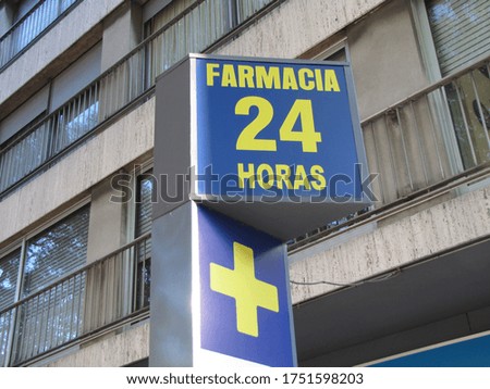 Poster with the sign, translation: "24 hour pharmacy" on the sidewalk. Street sign in blue with letters and yellow cross with sunlight illuminated apartment building background.

