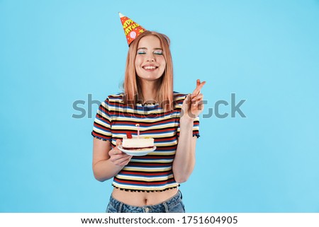 Image of cheerful girl in party cone holding cake and fingers crossed for good luck isolated over blue background