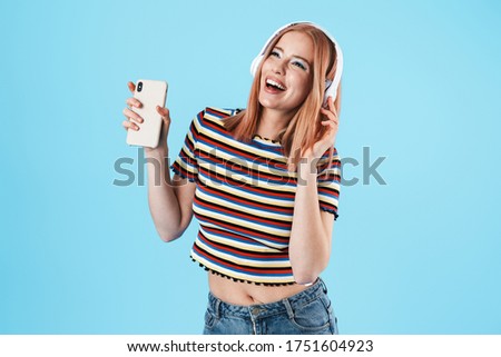 Image of cheerful caucasian girl using wireless headphones and cellphone isolated over blue background
