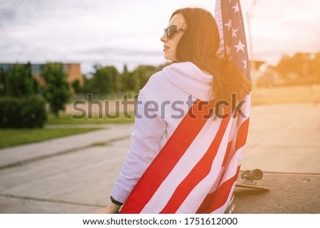 Beautiful patriotic vivacious young woman with the American flag held in her outstretched hands standing in the summer sunshine  in front of skate park