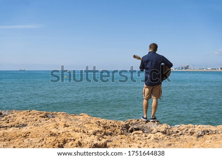 Young man with a printed shirt on his back playing a Spanish guitar standing in front of the sea with a port on the background