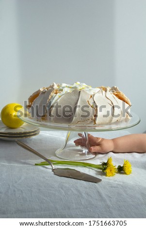 child's hand reaches for the meringue cake Pavlova with whipped cream and lemon on glass cake stand