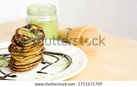 Matcha and banana pancakes with black chocolate, next to green matcha powder and Bamboo Matcha Whisk. Good looked and very tasty healthy breakfast on light background