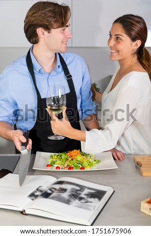 Handsome young man pointing at a recipe book with a knife and smiling at a young woman offering him a glass of white wine at the kitchen. Cooking at home and fun concept.