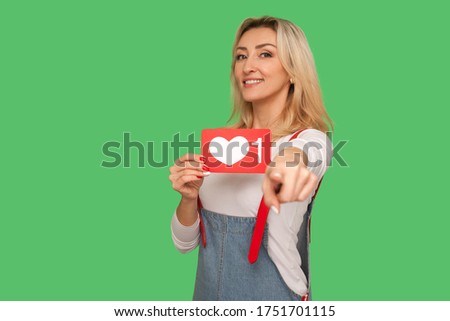 You push Like and follow social media blog! Portrait of lovely cheerful adult woman holding network heart icon and pointing to camera, showing emoji button. studio shot isolated on green background