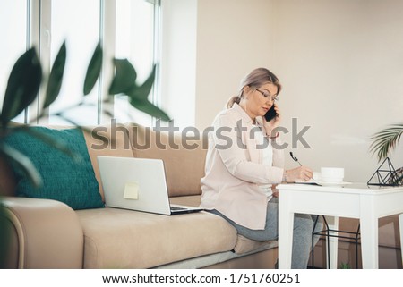 Senior caucasian woman with blonde hair and eyeglasses working from home at the pc while write something