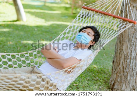 A young Asian man wearing a mask is sleeping happily on a hammock under a tree in a well-ventilated park, a new normal concept, keeping the distance safe during the COVID-19 epidemic.
