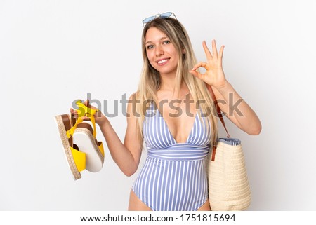 Young blonde woman holding summer sandals isolated on white background showing ok sign with fingers