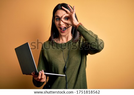 Young operator woman with blue eyes working on call center using computer laptop and headset with happy face smiling doing ok sign with hand on eye looking through fingers