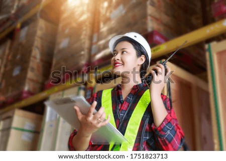 Warehouse logistic management business; young Asian woman wearing reflective safty vest and white helmet; Communicating via walky-talky in the warehouse factory as worker
