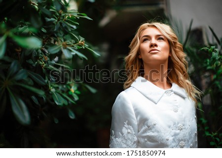 Portrait of an attractive blonde bride standing in a wedding dress on a background of greenery. The bride in the botanical green garden. Beautiful young woman with curly blond hair. Marriage.